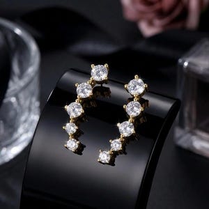 May include: A pair of gold-toned earrings with a curved design. Each earring features a series of graduated, round, clear gemstones. The earrings are displayed on a black, glossy surface, with a blurred background.