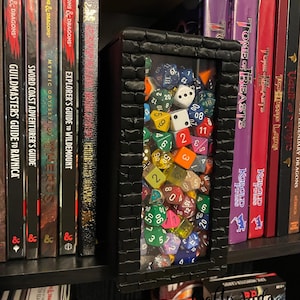 May include: A black wooden box with a glass front filled with colorful dice. The box is sitting on a shelf with books and game cards. The books are Dungeons & Dragons themed and the game cards are Monster Cards.