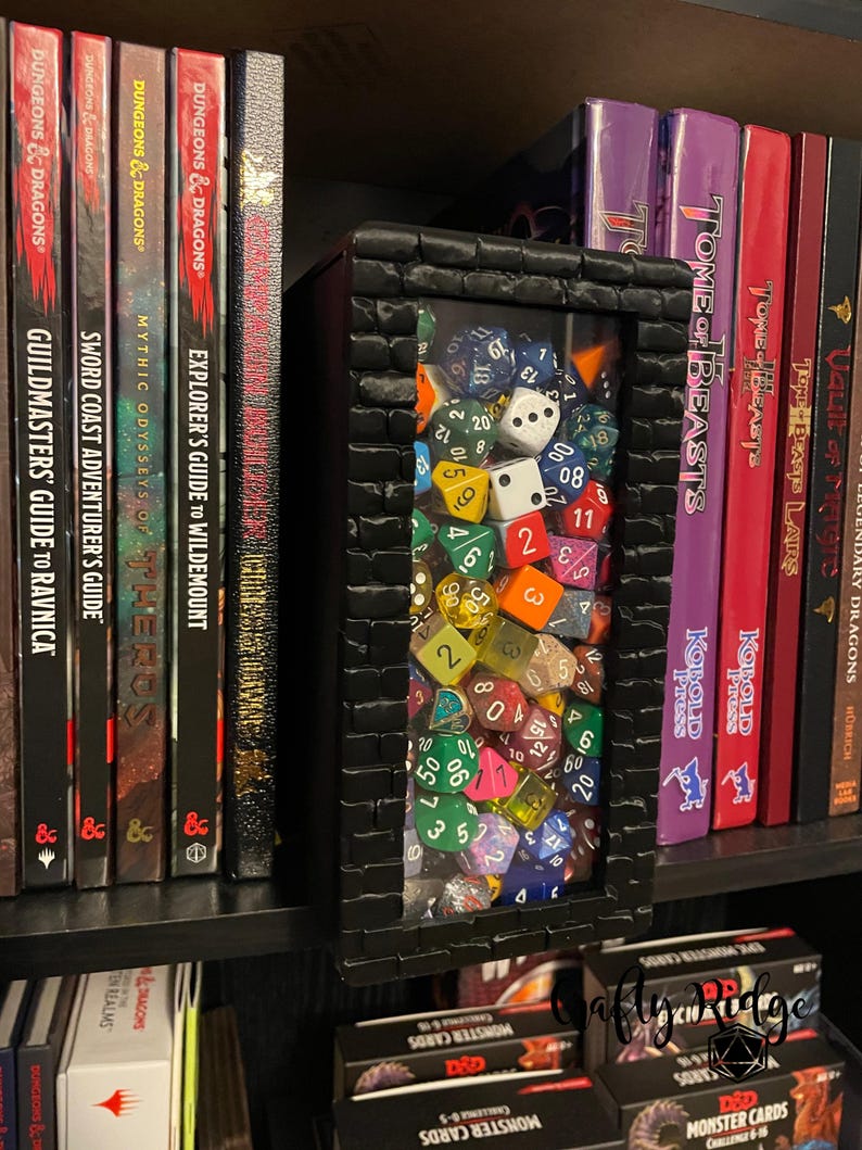 May include: A black wooden box with a glass front filled with colorful dice. The box is sitting on a shelf with books and game cards. The books are Dungeons & Dragons themed and the game cards are Monster Cards.