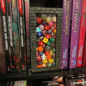 May include: A 3D printed bookend shaped like a brick wall with a window containing a collection of colorful dice. The bookend is on a bookshelf with several Dungeons and Dragons books and a Tome of Beasts book.
