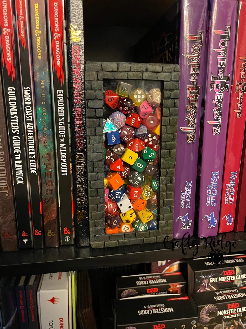 May include: A 3D printed bookend shaped like a brick wall with a window containing a collection of colorful dice. The bookend is on a bookshelf with several Dungeons and Dragons books and a Tome of Beasts book.