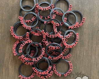 Può includere: Un mucchio di anelli di plastica nera con parole rosse stampate su di essi. Le parole includono "Haunted", "Frightened", "Inspired", "Poisoned", "Reckless", "Blinded", "Petrified", "Vicious", "Mocked", e altro ancora.