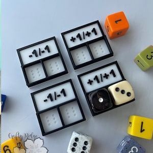 May include: Four black and white plastic game pieces with the text "-1/-1" and "+1/+1" on them.  The pieces are used to modify scores in a game.  There are also several dice in the image, including a white die with black dots, a black die with white dots, a yellow die with black dots, a light blue die with white dots, and an orange die with black dots.