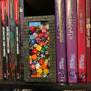 May include: A black and gray brick-shaped bookend with a clear window filled with colorful dice. The bookend is on a shelf with several books, including Dungeons & Dragons and Tome of Beasts.