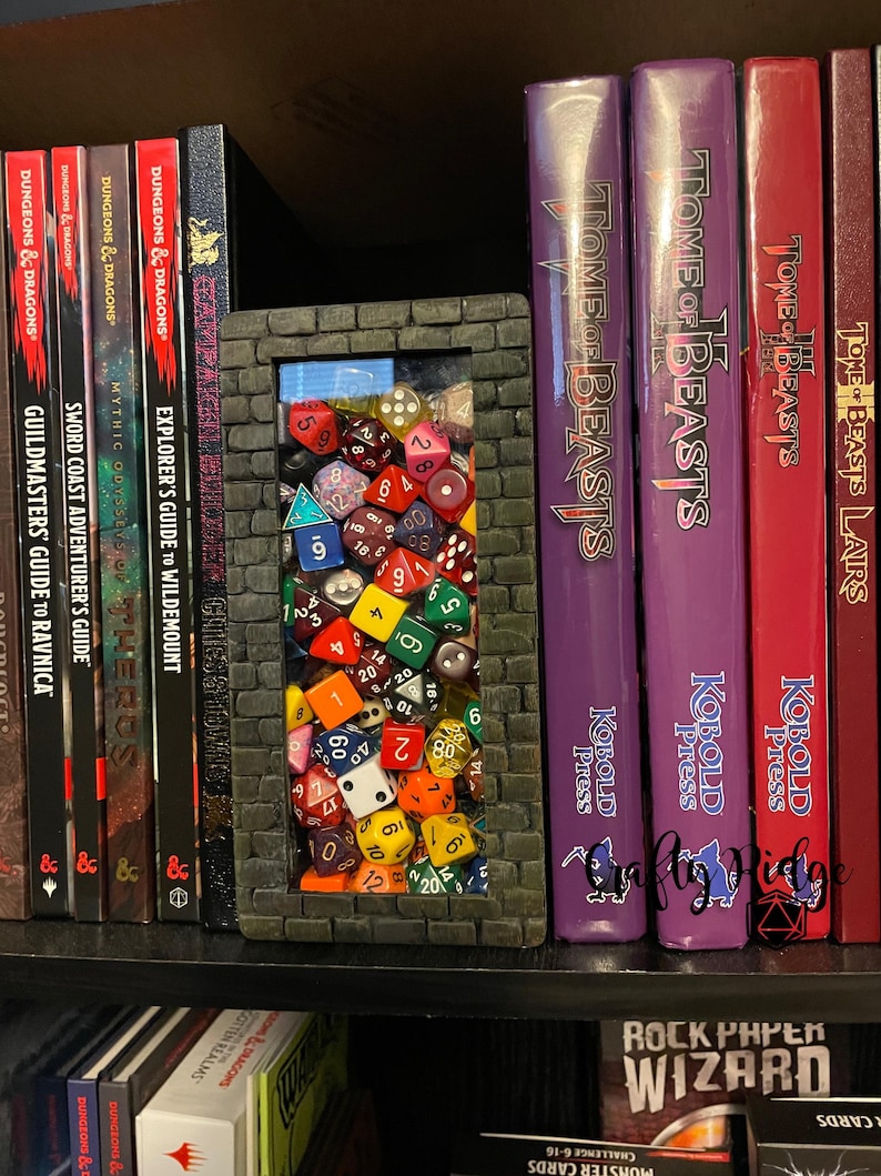 May include: A black and gray brick-shaped bookend with a clear window filled with colorful dice. The bookend is on a shelf with several books, including Dungeons & Dragons and Tome of Beasts.