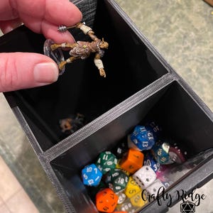 May include: A black plastic dice tower with two compartments. The top compartment holds a miniature figurine of a goblin. The bottom compartment is filled with colorful dice.