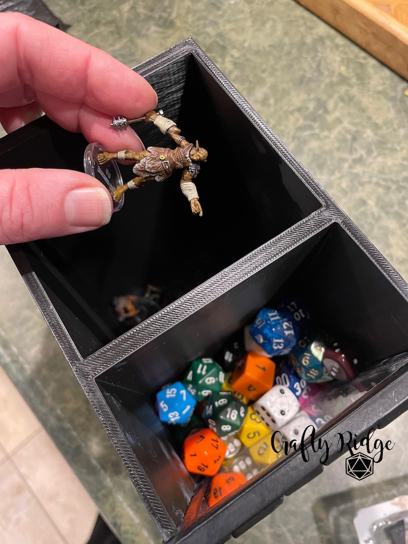 May include: A black plastic dice tower with two compartments. The top compartment holds a miniature figurine of a goblin. The bottom compartment is filled with colorful dice.