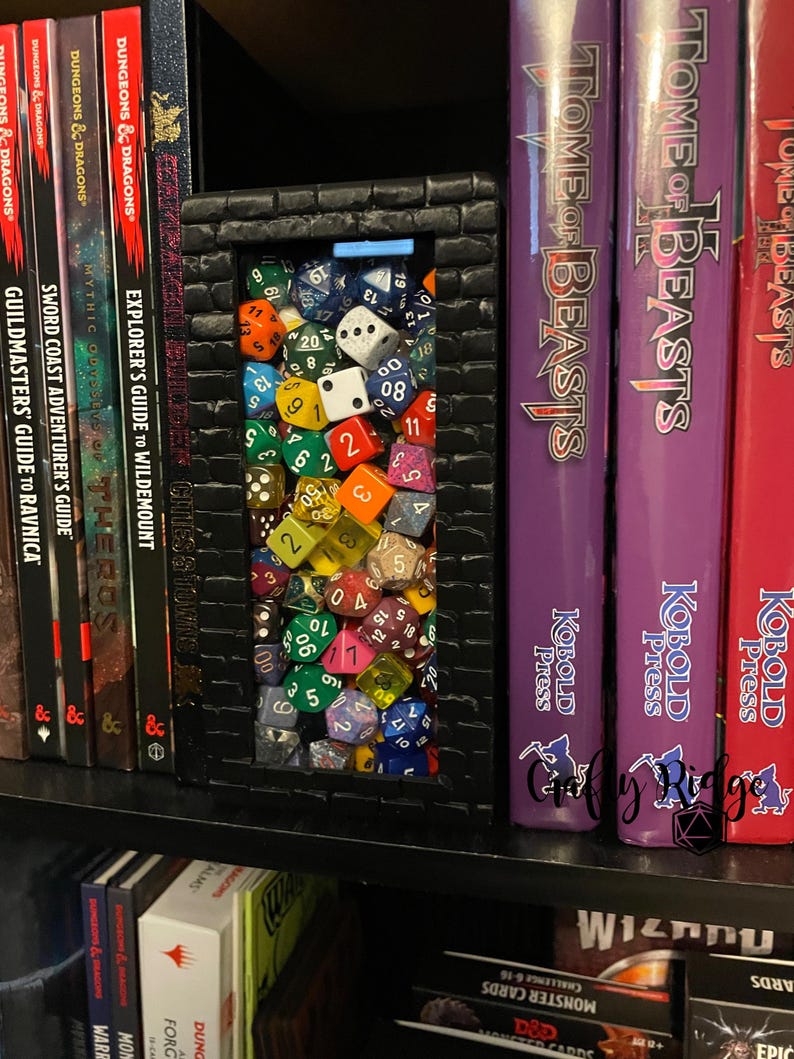 May include: A black brick-shaped bookend with a clear window filled with colorful dice. The bookend is on a bookshelf with several books, including "Tome of Beasts" and "Dungeon & Dragons" books.
