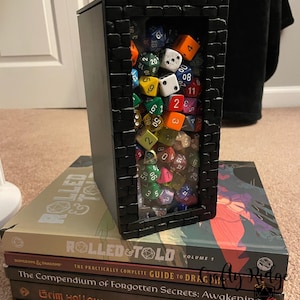 May include: A black box with a brick pattern frame, containing a large collection of colorful dice. The box is sitting on top of a stack of books with titles like "Rolled & Told", "The Compendium of Forgotten Secrets: Awakening", "Grim Hollow", and "Tales and Tomes from the Forbidden Library".