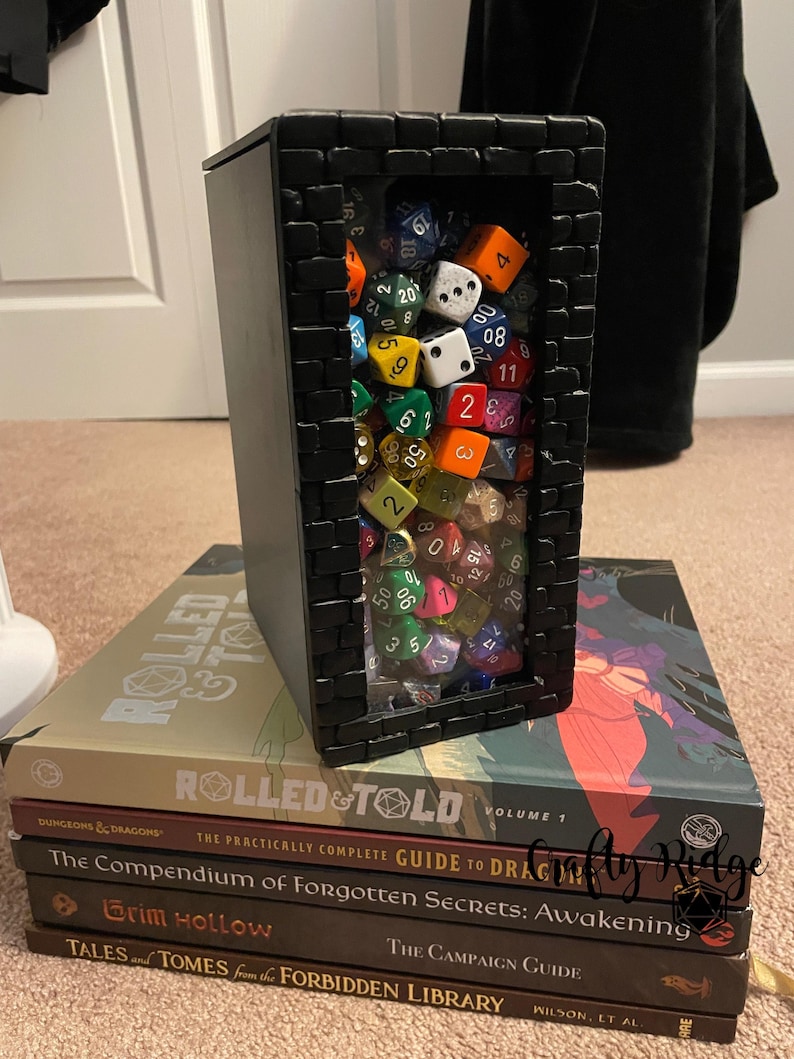 May include: A black box with a brick pattern frame, containing a large collection of colorful dice. The box is sitting on top of a stack of books with titles like "Rolled & Told", "The Compendium of Forgotten Secrets: Awakening", "Grim Hollow", and "Tales and Tomes from the Forbidden Library".