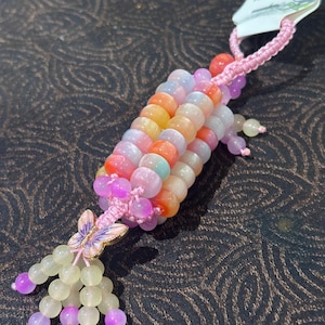 May include: A decorative charm featuring a cylindrical cluster of translucent, multi-coloured beads in shades of orange, pink, and white. A small metal butterfly charm with pink and purple enamel accents hangs from the bottom, finished with a pink cord.
