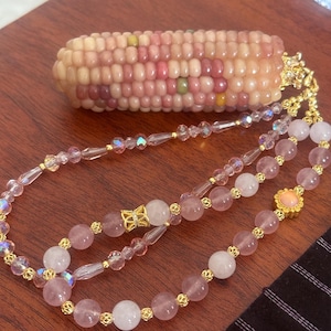 May include: A decorative corn-shaped pendant crafted from small, multi-coloured beads in shades of pink, peach, and green, attached to a long beaded necklace featuring translucent pink crystals, round rose quartz beads, and small gold-toned spacers.