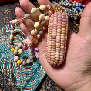 Könnte beinhalten: Eine einzigartige Halskette mit einem Anhänger in Form eines Maiskolbens mit rosa, gelben und weißen Körnern. Die Halskette enthält einen Perlenstrang mit verschiedenen pastellfarbenen Perlen. Im Hintergrund befinden sich weiterer Schmuck und ein gemusterter Stoff.
