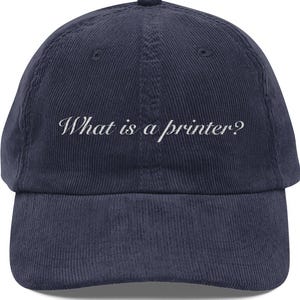 May include: Navy blue corduroy baseball cap with the text "What is a printer?" embroidered in white. The cap has a curved brim and a button on top. A casual, everyday accessory.