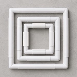 Può includere: Tre telai da ricamo in plastica bianca annidati l'uno nell'altro, creando un design quadrato-in-quadrato. I telai sono costituiti da sezioni dritte e curve collegate, con un'estetica semplice e pulita. Lo sfondo è una superficie grigia chiara e uniforme.