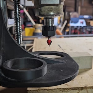 Può includere: Primo piano di una macchina CNC con base nera e punta rossa appuntita. La macchina è posizionata sopra una superficie di legno, con un piccolo blocco di legno nelle vicinanze. Lo sfondo mostra un'officina.