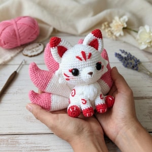 May include: A handmade crochet fox doll with white, pink, and red accents. The fox has large black eyes, a fluffy white chest, and a tail with pink and white stripes. The doll is held in two hands.