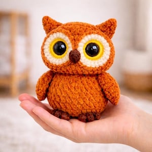 May include: A handmade, orange crochet owl with large black eyes, yellow and white eye accents, and a brown beak. The owl has small ear tufts and is held in a person's hand. The owl is a decorative item.
