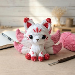 May include: A handmade crochet fox doll with white, pink, and red accents. The fox has large black eyes, a small nose, and a fluffy tail. The fox is sitting on a light-colored surface. A pink ball of yarn and a crochet hook are also visible.
