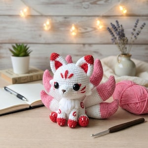 May include: A handmade crochet fox doll with white, pink, and red accents. The fox has large black eyes, a fluffy white chest, and a tail with multiple pink and white sections. The fox is sitting on a wooden surface.