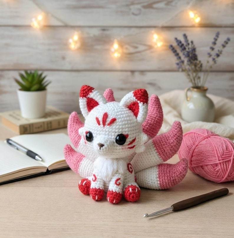 May include: A handmade crochet fox doll with white, pink, and red accents. The fox has large black eyes, a fluffy white chest, and a tail with multiple pink and white sections. The fox is sitting on a wooden surface.