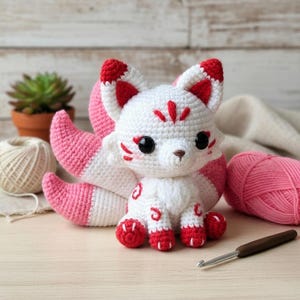 May include: A handmade crochet fox with white and pink details. The fox has large black eyes, red accents on its ears and paws, and a fluffy tail. The fox is sitting on a light-colored surface.