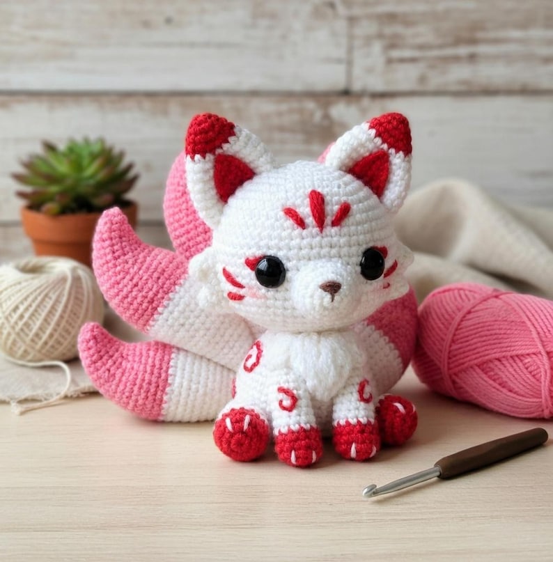 May include: A handmade crochet fox with white and pink details. The fox has large black eyes, red accents on its ears and paws, and a fluffy tail. The fox is sitting on a light-colored surface.