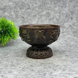May include: A dark bronze-toned metal bowl with a pedestal base, featuring intricate raised relief carvings of floral and knot-like motifs around the exterior. The bowl has a wide, open rim and a rounded body that tapers down to a fluted base.
