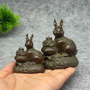 May include: Two bronze-colored rabbit figurines, each sitting beside a small pot filled with coins. The rabbits have textured fur details and are positioned on a base decorated with a woven pattern and circular coin motifs.
