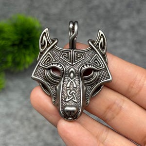 Könnte beinhalten: Ein silberfarbener Metallanhänger in Form eines stilisierten Wolfskopfes. Die Oberfläche weist komplexe gravierte geometrische Muster, eine texturierte Punktierung und ein zentrales Knotenmuster auf dem Fang auf. Eine kleine Öse oben dient zur Aufhängung.