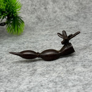 Pode incluir: Uma escultura estilizada marrom escuro de uma libélula. A escultura tem um corpo longo e curvo com uma extremidade pontiaguda e uma seção média bulbosa. As asas da libélula estão abertas e a escultura é feita de um material escuro, possivelmente metálico.