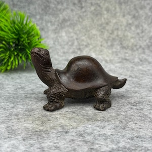 Op de afbeelding: Een kleine, gedetailleerde schildpadfiguur in bronskleur. De sculptuur heeft een getextureerd schild, gedetailleerde poten en een geconcentreerde uitdrukking. Het beeldje is een decoratief item, waarschijnlijk voor weergave.