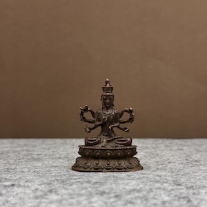 Op de afbeelding: Een kleine, bronskleurige beeld van een veelarmige godheid, zittend op een lotusbloem basis. De figuur draagt een kroon en heeft meerdere armen die verschillende objecten vasthouden. De achtergrond is neutraal.