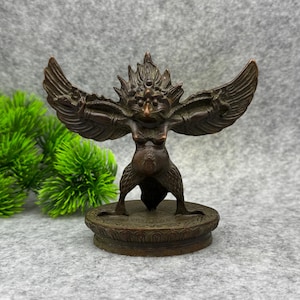 Può includere: Una statuetta in metallo color bronzo raffigurante una creatura mitica con ali spiegate, testa di uccello e petto prominente. La figura poggia su una base circolare con intricati motivi geometrici e un design a petali di loto sul bordo.