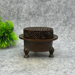Puede incluir: Un quemador de incienso de color bronce con una parte superior estampada y una base en forma de cuenco. El quemador tiene tres patas pequeñas y redondeadas y un diseño decorativo calado en la parte superior. El diseño es ornamentado y detallado.