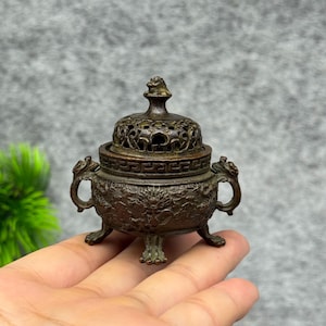 May include: A small, dark bronze-toned metal incense burner featuring an intricate lid with a finial, two side handles shaped like mythical creatures, and three legs. The body is decorated with detailed relief carvings of dragons and geometric patterns.