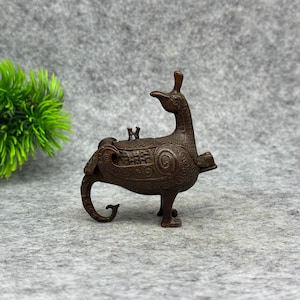 May include: A dark bronze-colored metal vessel shaped like a mythical bird with a long neck, a curved tail, and intricate swirling patterns engraved across its body. The object features a small lid on its back and stands on two sturdy legs.