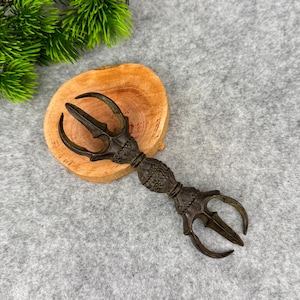 May include: A bronze-coloured vajra, a ritual object, rests on a wooden slice. The vajra features a three-pronged design at each end and a detailed, textured handle. Green foliage is visible in the upper left corner, and the background is a grey fabric.