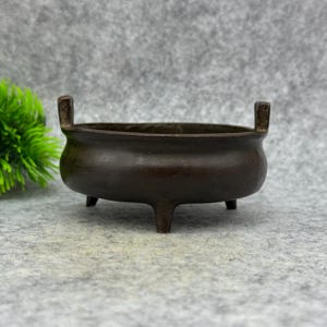 May include: A bronze incense burner with a rounded bowl shape and three small legs. The burner has two small, rectangular handles on either side of the rim. The design is simple and elegant.