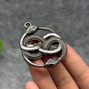 Könnte beinhalten: Ein kreisförmiger silberfarbener Metallanhänger, der zwei ineinander verschlungene Schlangen zeigt, die eine Unendlichkeitsschleife bilden. Die Schlangen haben detaillierte Schuppenstrukturen auf ihren Körpern und ausgeprägte Köpfe mit sichtbaren Augen. Oben befindet sich eine kleine Öse.