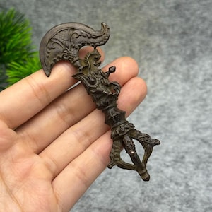 May include: Bronze-colored, ornate ritual object with a curved blade and intricate dragon-like details. The handle features a three-pronged design. The object is held in a hand, showcasing its size and craftsmanship. The overall design suggests a ceremonial or decorative purpose.
