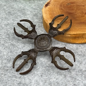 May include: A bronze-coloured, four-pronged ritual object with a central circular design. Each prong curves outwards, ending in a three-pronged tip. The object is likely used for religious or ceremonial purposes.