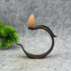 May include: A dark brown incense holder with a curved design, holding a tan incense cone. The holder is made from a wood-like material and has a smooth finish. The incense cone is a light brown colour and is placed on top of the holder.