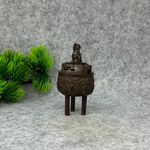 May include: A bronze-colored incense burner with a detailed design. The burner has a lid with a small statue on top and stands on three legs. The body of the burner is decorated with intricate patterns.