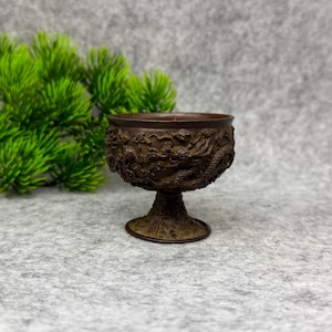 Könnte beinhalten: Ein bronzefarbenes, verziertes Kelchglas mit einem detaillierten Drachenmotiv, das um die Schale geschnitzt ist. Das Kelchglas hat einen Sockel und einen breiten Rand. Der Hintergrund ist ein neutrales Grau, mit etwas grünem Laub auf der linken Seite. Es ist etwa 8 cm hoch.