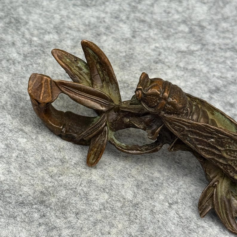 以下が含まれることがあります： ブロンズ色の昆虫型の装飾品。このオブジェクトは、詳細な翼と湾曲したハンドルを備えています。昆虫はセミまたは同様の種であるようです。このオブジェクトは、風化したアンティークの外観をしています。