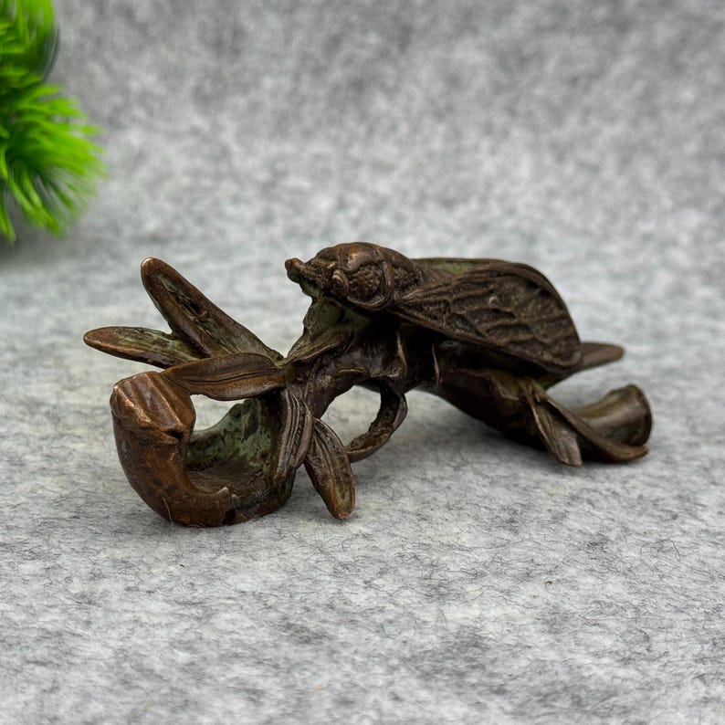 以下が含まれることがあります： ブロンズ色の彫刻で、様式化された植物にとまっているセミを表しています。昆虫は詳細な羽と体を持っており、植物は湾曲した葉を持っています。彫刻はおそらく金属製で、風化した外観をしています。