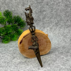 Może przedstawiać: Ozdobny, brązowy rytualny sztylet z głową konia na górze i głową orła na dole. Sztylet spoczywa na drewnianym plastrze na szarym tle. Złożony wzór sugeruje przedmiot ceremonialny lub religijny.