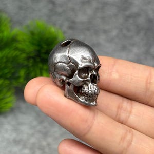 May include: A small, three-dimensional metallic skull bead with a dark, weathered finish. The skull features detailed anatomical contours, including visible suture lines on the cranium, deep eye sockets, a nasal cavity, and teeth.