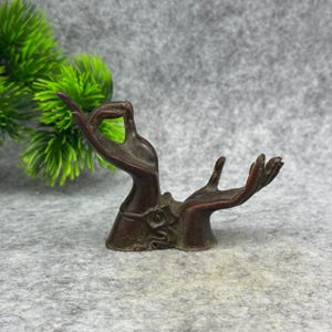 Può includere: Una scultura color bronzo raffigurante due mani in un gesto di offerta o benedizione. Le mani sono dettagliate con linee e curve visibili e la scultura ha un aspetto invecchiato. La base presenta un design ornato.
